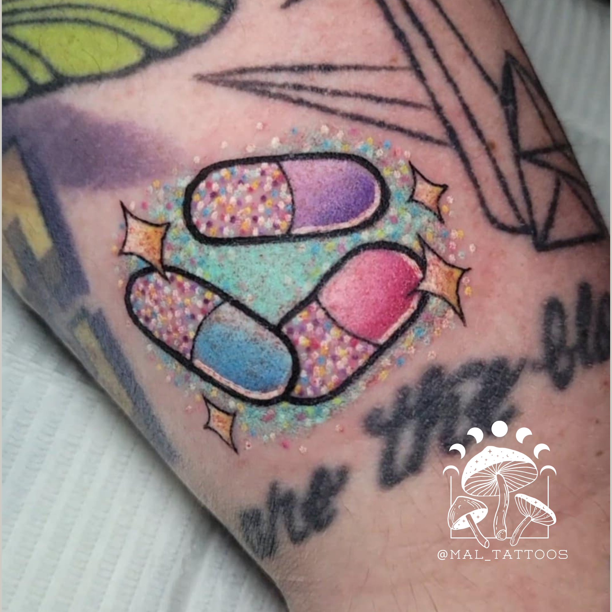 Colorful胶囊 tattoo with glitter on a person's arm, featuring the artist's logo in the corner.