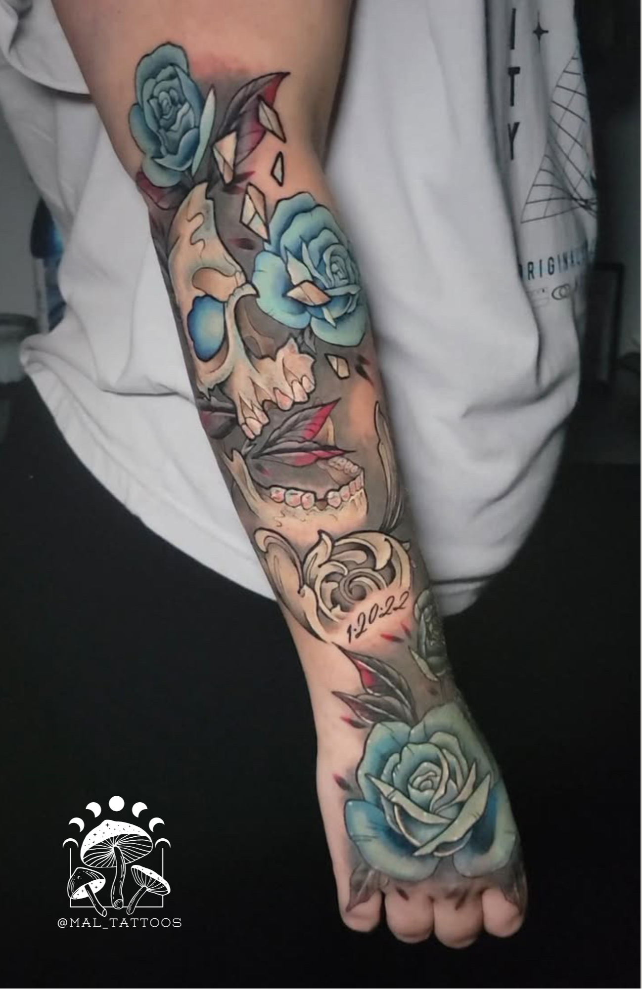 Detailed tattoo design on a person's arm with flowers and skull and other elements, featuring the Mal Tattooos logo.
